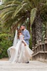 Одружена пара в одязі стоїть на дерев'яному пішохідному мосту з перилами, цілуючись біля зелених долонь і рослин в саду в літній день — стокове фото