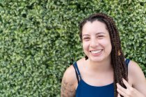 Positive Hispanic female with tattoo and long black braided hair smiling and looking at camera while standing near green plants — Stock Photo