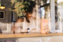 Через вікно радісної молодої етнічної жінки з темним афро волоссям у модному вбранні та справжніми бездротовими навушниками, що посміхаються щасливо, маючи відео розмову на смартфоні в сучасному кафе — стокове фото