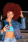 Весела жінка з африканською зачіскою, одягнена в модний денімний одяг, в якому повідомляють по мобільному телефону, стоячи біля паркану на вулиці увечері. — стокове фото