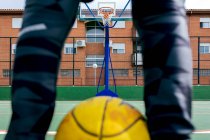 Анонимный спортсмен в спортивной одежде, стоящий на спортивной площадке с желтым мячом и баскетбольным кольцом во время игры на улице — стоковое фото