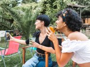 Encantada multirracial mejores amigas con vasos de cócteles escalofriantes en el parque de verano y tomar selfie en el teléfono inteligente - foto de stock