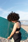 Вид сбоку на молодую женщину-серфингистку в гидрокостюме с доской для серфинга, стоящую, отводя взгляд на берег, уносимый волнами моря — стоковое фото