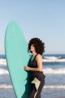 Vista lateral de la joven surfista en traje de neopreno con tabla de surf de pie mirando hacia otro lado en la orilla del mar bañada por el mar ondulante - foto de stock