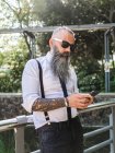 Confident bearded hipster in stylish outfit and sunglasses text messaging on cellphone while standing near metal fence on sunny street — Stock Photo