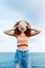 Unrecognizable female with long ginger curls covering face with hat on coast of blue sea — Stock Photo