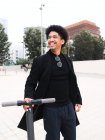 Moda jovem afro-americano cara com cabelo encaracolado escuro em roupa elegante em pé na praça da cidade e olhando para longe depois de andar de scooter — Fotografia de Stock