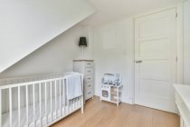 Weißes Kinderbett mit Handtuch im hellen, stilvollen Schlafzimmer mit Lampe auf der Kommode und Spielzeug in der Nähe geschlossener Tür — Stockfoto