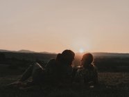 Casal de hippies com óculos de sol deitados em um prado gramado na natureza enquanto observava o pôr do sol entre as montanhas — Fotografia de Stock