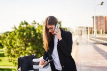 Concentrated businesswoman with brown hair in stylish clothes standing near concrete fence with cup of takeaway coffee and bag and whiting notes in notebook on street in sunny day while talking on the mobile — Stock Photo