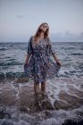 Delighted female with long hair in trendy dress standing with closed eyes on seashore in summer evening — Stock Photo