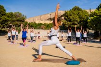 Side view of unrecognizable women in white clothes standing with raised arms and performing Virabhadrasana B during yoga lesson in park in sunny day — Stock Photo