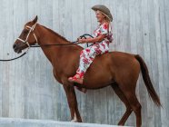 Visão lateral corpo inteiro de fêmea sênior sorridente em chapéu de cowboy segurando rédea no garanhão castanho — Fotografia de Stock