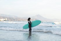 Vue latérale de jeune surfeuse réfléchie en combinaison avec planche de surf debout regardant loin sur le bord de mer lavé par la mer ondulante — Photo de stock