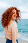 Visão traseira da fêmea encantada com cabelos longos encaracolados de gengibre olhando sobre o ombro para a câmera contra o mar azul — Fotografia de Stock