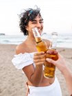 Радостная этническая женщина, звенящая бутылка пива с другом урожая, наслаждаясь летним вечером на песчаном пляже — стоковое фото