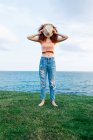 Full body of unrecognizable barefoot female with long ginger curls covering face with hat on grassy coast of blue sea — Stock Photo
