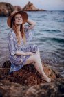 Jovem encantadora em vestido de verão e chapéu sentado na costa rochosa enquanto olha para longe na noite de verão — Fotografia de Stock
