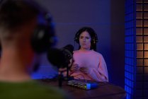 Молода жінка-радіоведуча в повсякденному одязі і навушники, сидячи за столом з мікрофоном і спілкуючись з анонімним колегою врожаю під час подкасту в студії — стокове фото