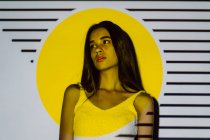 Созерцательная молодая латиноамериканка с макияжем и длинными волосами, смотрящая в желтый проектор света с полосками — стоковое фото