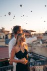 Liebender Mann umarmt Frau von hinten und zeigt auf Dachterrasse mit Heißluftballons am Abendhimmel in Kappadokien Türkei — Stockfoto
