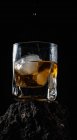Капли виски, падающие на кубики льда, подаются в хрустальном стекле, размещенном на грубой поверхности на черном фоне — стоковое фото