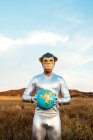 Anonymer Typ im silbernen Latex-Anzug mit geometrischer Affenmaske, der in die Kamera schaut und einen Globus in der Natur hält — Stockfoto