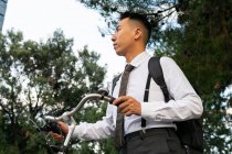 Side view of dreamy young ethnic male office worker with backpack and bike looking away against urban building and trees — Stock Photo