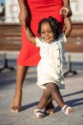 Fröhliches afroamerikanisches Mädchen in hellem Kleid und High Heels, das die Hände einer nicht wiederzuerkennenden Mutter hält, während es an sonnigen Tagen auf der Straße spaziert — Stockfoto