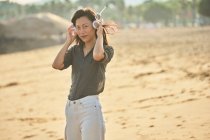 Vue latérale de paisible femme asiatique réfléchie regardant la caméra écouter la chanson des écouteurs sans fil tout en marchant sur la rive sablonneuse — Photo de stock