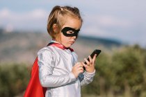 Vista lateral da menina auto-assegurada no traje de super-herói máscara de olho com capa navegando no celular — Fotografia de Stock