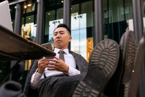 Внимательный молодой этнический бизнесмен сидит со скрещенными ногами, просматривая интернет по мобильному телефону за столом в кафетерии в городе — стоковое фото