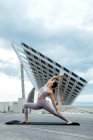 Corpo completo di donna sportiva in activewear praticare Viparita Virabhadrasana postura durante l'allenamento su strada vicino al pannello solare contro cielo nuvoloso — Foto stock