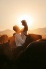 Vue latérale d'une femme heureuse admirant le coucher du soleil sur les montagnes tout en étant élevée par un homme aimant parmi des chevaux calmes dans un champ de Turquie — Photo de stock