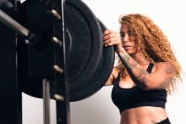 Femme de sport forte avec les cheveux bouclés en vêtements de sport debout et mettre la plaque de poids sur haltère pendant l'entraînement dans la salle de gym — Photo de stock