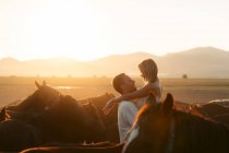 Vista lateral de la mujer feliz admirando la puesta de sol sobre las montañas mientras es criado por el hombre amoroso entre caballos tranquilos en el campo de Turquía - foto de stock
