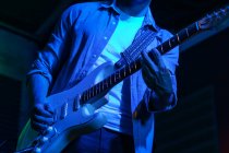 Crop anonymous male guitarist playing electric guitar in club with neon blue lights — Stock Photo