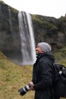 Seitenansicht des Fotografen männlichen Touristen in warmer Oberbekleidung und Rucksack bewundern erstaunliche Aussicht auf den Seljalandsfoss Wasserfall fließt durch felsige Klippe in Teich — Stockfoto