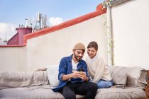 Усміхнена молода жінка сидить поруч з бородатим хлопцем і переглядає смартфон на дивані на терасі — стокове фото