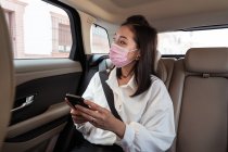 Asian female passenger in protective mask in formal clothes browsing cellphone while sitting on backseat in taxi and looking away with interest — Stock Photo