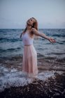 Attraente giovane femmina in abiti estivi alla moda in piedi con le braccia alzate e gli occhi chiusi in acqua di mare in sera — Foto stock