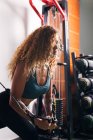 Сильная спортсменка с королевскими вьющимися волосами в спортивной одежде делает упражнения на кабельном кроссовере в тренажерном зале днем — стоковое фото