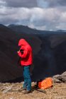 Vista lateral del fotógrafo masculino en ropa de abrigo de pie en la parte superior del acantilado rocoso cerca del volcán activo Fagradalsfjall con lava negra en Islandia durante el día - foto de stock