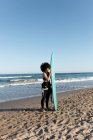Side view of young female surfer in wetsuit with surfboard standing looking away on seashore washed by waving sea — Stock Photo