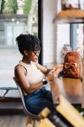 Vista laterale di fiduciosa giovane afro-americana millenaria con capelli ricci scuri in abito alla moda e occhiali da sole seduti a tavola in caffè moderno e messaggistica su smartphone — Foto stock