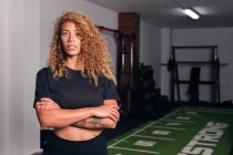 Selbstbewusste hispanische Sportlerin mit langen lockigen Haaren in Activwear steht mit verschränkten Armen im Fitnessstudio und blickt in die Kamera — Stockfoto