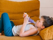 Вид збоку маленького хлопчика з астмою, який використовує інгалятор, лежачи на дивані вдома — стокове фото
