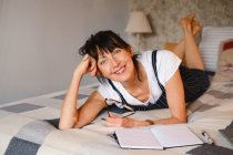 Positive middle age female leaning on hand while lying on bed with opened notepad and eyeglasses and looking at camera — Stock Photo