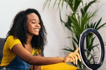 Afroamerikanerin in Jeans lächelt auf Sofa und berührt Smartphone an glühender Ringlampe — Stockfoto