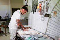 Боковой вид молодого татуированного пекаря в белой рубашке поло и очках, разминающего тесто руками, стоя у металлического прилавка на кухне современной пекарни — стоковое фото